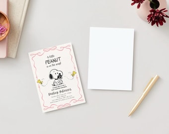 May include: A baby shower invitation with a Snoopy illustration and the text "A little peanut is on the way!". The invitation is light pink with a decorative border. A blank white card, a gold pen, and flowers are also on the white surface.