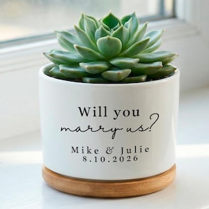 May include: A white ceramic planter with a succulent plant. The planter has the text "Will you marry us?" along with names and a date. The planter sits on a round wooden base, perfect for a proposal.