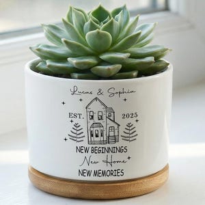 Pode incluir: Um vaso de cerâmica branco com uma planta suculenta. O vaso tem uma base de madeira e apresenta uma ilustração de uma casa com o texto "Lucas & Sophia, New Beginnings, New Home, New Memories, Est. 2025."