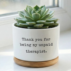 May include: A white ceramic plant pot with a green succulent. The pot has the text "Thank you for being my unpaid therapist." It sits on a round, wooden base. The plant is a vibrant green colour.