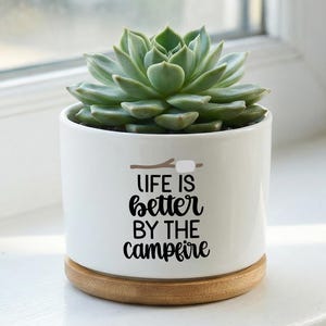 May include: A white ceramic plant pot with a succulent. The pot has the phrase "LIFE IS better BY THE campfire" in black script. It sits on a round, light brown wooden base. The succulent has green leaves.