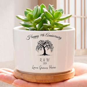May include: White ceramic planter with a green succulent plant, resting on a wooden base. The planter is inscribed with "Happy 9th Anniversary", a tree illustration, and "Love Grows Here". A thoughtful gift.
