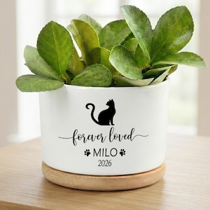 May include: A white ceramic planter with a wooden base, holding green leafy plants. The planter features a black silhouette of a cat, the words "forever loved", the name "MILO", and the year "2026".