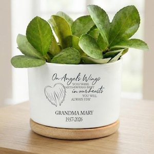 Op de afbeelding: Een witte keramische plantenpot met een houten basis, gevuld met groene bladplanten. De pot is voorzien van de tekst "On Angel's Wings" en "Grandma Mary 1937-2026". Een hart met vleugels is ook op de pot gedrukt.