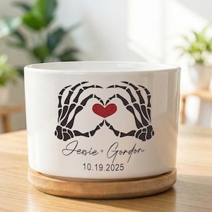 May include: White ceramic planter with a black skeleton hand heart design and a red heart in the center. The names "Jessie + Gordon" and the date "10.19.2025" are written below the heart. The planter sits on a wooden coaster.