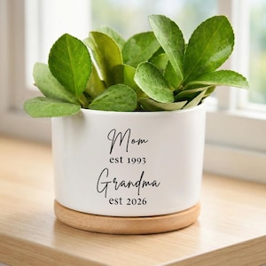 Puede incluir: Una maceta de cerámica blanca con las palabras "Mom est 1993 Grandma est 2026" en escritura negra. La maceta contiene una planta verde y descansa sobre una base de madera. La maceta está sobre una superficie de madera cerca de una ventana.