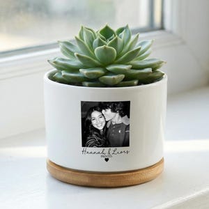 Peut inclure: Un pot de fleurs en céramique blanche avec une photo en noir et blanc d'un couple et le texte "Haanah & Liors Prints" avec un cœur. Le pot contient une plante succulente verte et repose sur une base en bois.