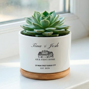 Könnte beinhalten: Weißer Keramik-Pflanztopf mit einer Sukkulente. Der Topf hat einen Holzsockel und trägt die Aufschrift "Tina & Josh", eine Hausillustration, "OUR FIRST HOME", Adresse und "EST 2026".
