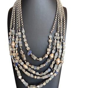 May include: A multi-strand necklace displayed on a black mannequin. The necklace features silver-toned chains and multiple strands of square and round beads in shades of gray, silver, and blue. The necklace has a layered design.