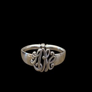 May include: Silver-toned hinged bangle bracelet with an ornate monogram design. The monogram features the letters "AMK" in a flowing, stylized script. The bracelet has a simple, polished band and a clasp closure.