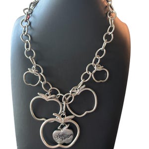 May include: A silver-toned chain necklace featuring multiple open apple-shaped charms. The central charm is a solid apple pendant with the text Apple Bottoms engraved on its surface. The necklace is constructed from circular metal links.
