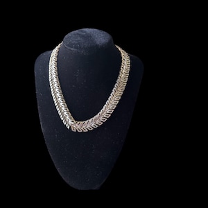 May include: A gold-toned necklace with a repeating leaf design is displayed on a black velvet bust. The necklace has a substantial weight and a polished finish, reflecting light. The background is a solid black, emphasizing the jewelry.