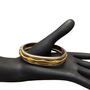 May include: A two-tone bangle bracelet with a gold-colored center band and a silver-colored outer band. The bracelet has a square shape with rounded edges. The gold band has a wavy texture.