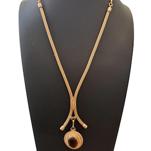 May include: A gold-toned necklace with a mesh chain and a pendant. The pendant features a dark brown gemstone surrounded by a decorative gold frame. The necklace is displayed on a black mannequin.