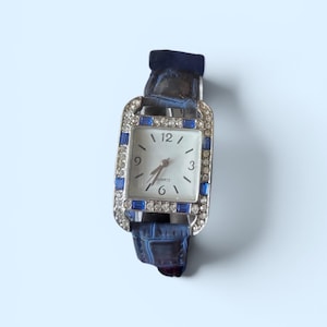 May include: A rectangular wristwatch with a white face, black numerals at 3, 6, 9, and 12, and the word QUARTZ printed below the center. The silver-tone bezel is encrusted with clear crystals and blue baguette stones. It has a dark blue leather strap.