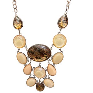 May include: A silver-toned necklace featuring a cascade of oval and teardrop-shaped stones in shades of cream, peach, and brown. The central stone is an oval with a marbled pattern. The necklace has a chain closure.