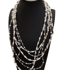 May include: A multi-strand necklace displayed on a black mannequin. The necklace features alternating white and silver-colored beads. The strands cascade down, creating a layered effect. The beads are small and evenly spaced, giving the necklace a delicate appearance.