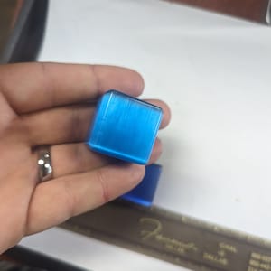 May include: A small, square, blue, polished, gemstone with a striated pattern.