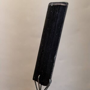 May include: A black, rectangular tool with a pointed tip and a set of metal hooks at the base. The tool is made of a smooth, black material and has a slightly textured surface.