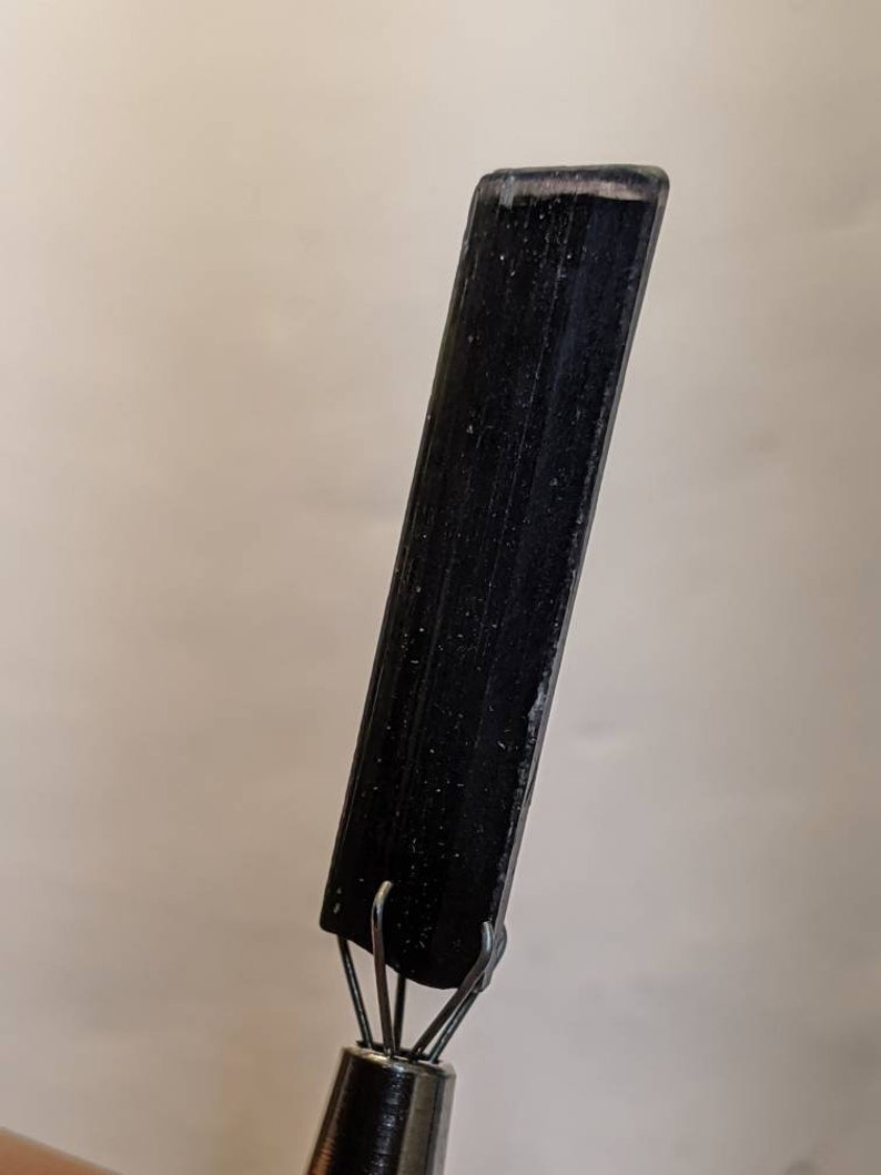 May include: A black, rectangular tool with a pointed tip and a set of metal hooks at the base. The tool is made of a smooth, black material and has a slightly textured surface.