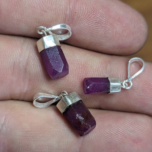 May include: Three silver pendants with purple gemstone crystals. Each pendant features a silver bail for attaching to a chain or cord.