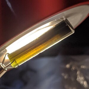 May include: A close-up of a clear glass tube filled with a thick, golden-yellow liquid. The liquid has a slight green tint and is suspended in the tube by a metal clip.