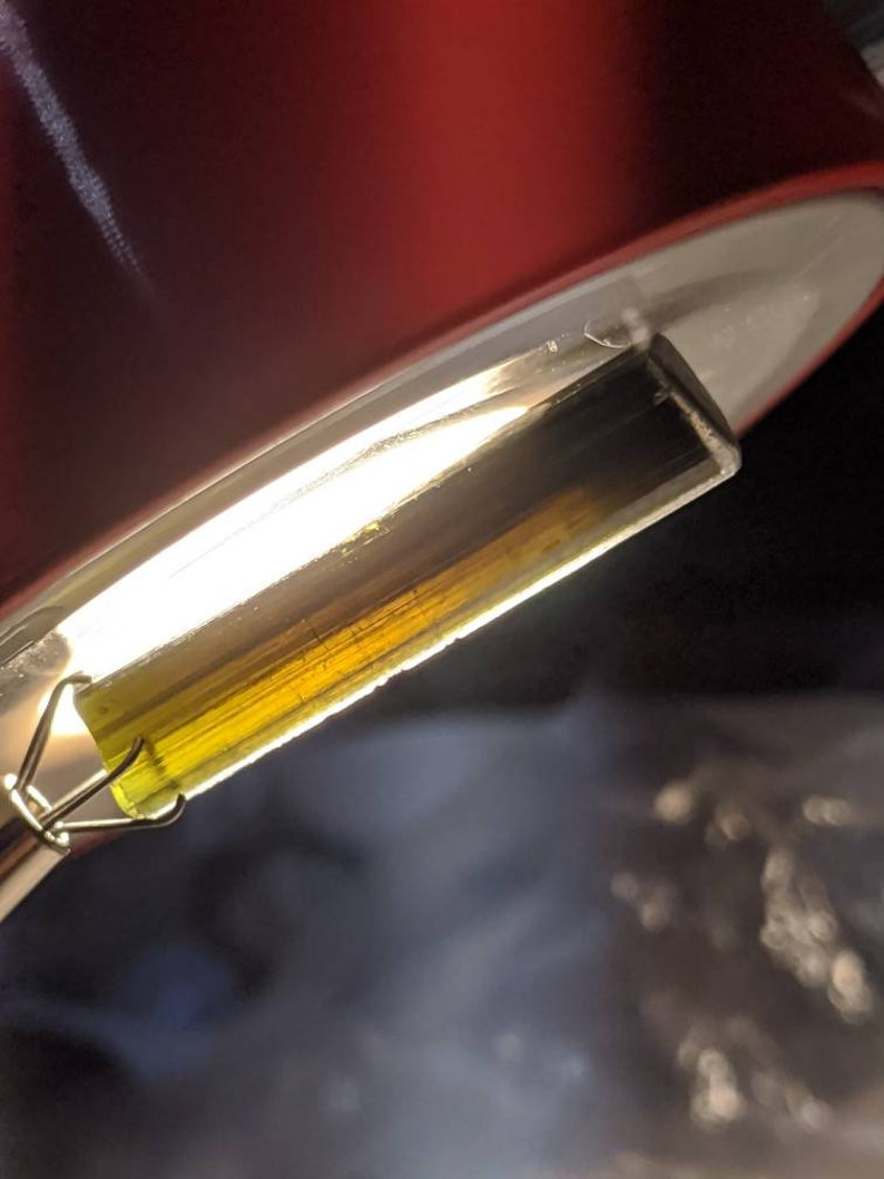 May include: A close-up of a clear glass tube filled with a thick, golden-yellow liquid. The liquid has a slight green tint and is suspended in the tube by a metal clip.