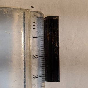 May include: A black metal object measuring approximately 2.5 cm in length. The object is rectangular in shape and has a slightly rounded edge.
