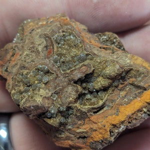 May include: A rough, natural rock formation with a mix of colors. The rock features a base of brown and reddish-brown hues, with streaks of bright orange and patches of small, sparkling crystals.