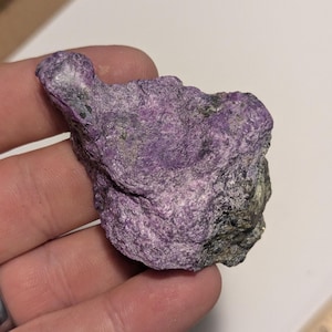 May include: A rough, purple and gray rock with a slightly metallic sheen. The rock is about the size of a person's hand.