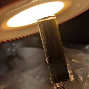 May include: A close-up of a dark green, cylindrical, crystal-like object held in a metal clamp. The object is illuminated from above by a bright light.