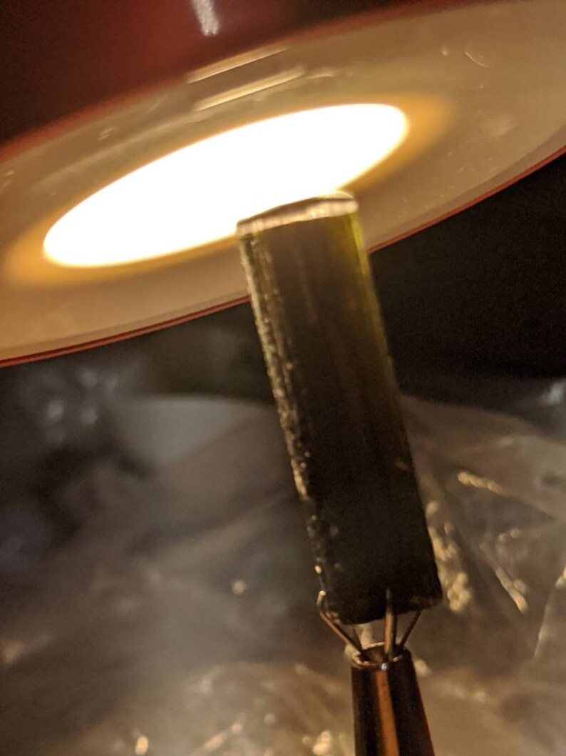 May include: A close-up of a dark green, cylindrical, crystal-like object held in a metal clamp. The object is illuminated from above by a bright light.