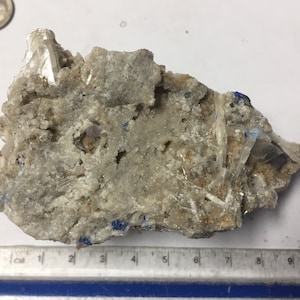 May include: A rough, irregularly shaped mineral specimen with a light gray and tan matrix. Clear, crystalline structures are visible throughout, with small patches of blue. A ruler with inch markings is at the bottom.