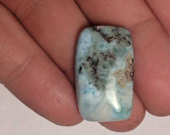 May include: A light blue and white cabochon gemstone with black and silver inclusions. The gemstone is cut in a rectangular shape.