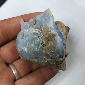 May include: A light blue opal crystal with a rough, natural texture. The crystal is partially embedded in a brown, rocky matrix.