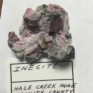 May include: A pink and grey inesite mineral specimen from Hale Creek, Trinity County, California. The label also includes the name of the seller, Claude Huber Minerals.