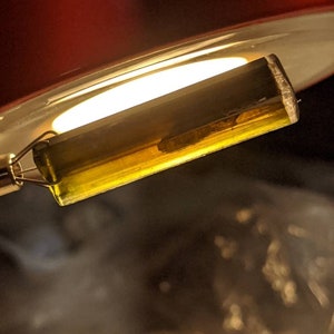 May include: A close-up of a clear glass tube with a yellow liquid inside. The tube is attached to a metal wire and is being held over a pot of boiling water.