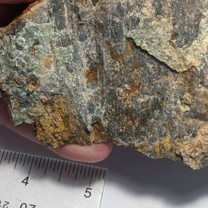 Puede incluir: Primer plano de una roca gris oscura con marcas verdes y marrones. La roca mide aproximadamente 12,5 cm de largo y se sostiene en una mano.
