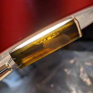 May include: A close-up of a green and gold gemstone with a white inclusion. The gemstone is held in a metal clamp.