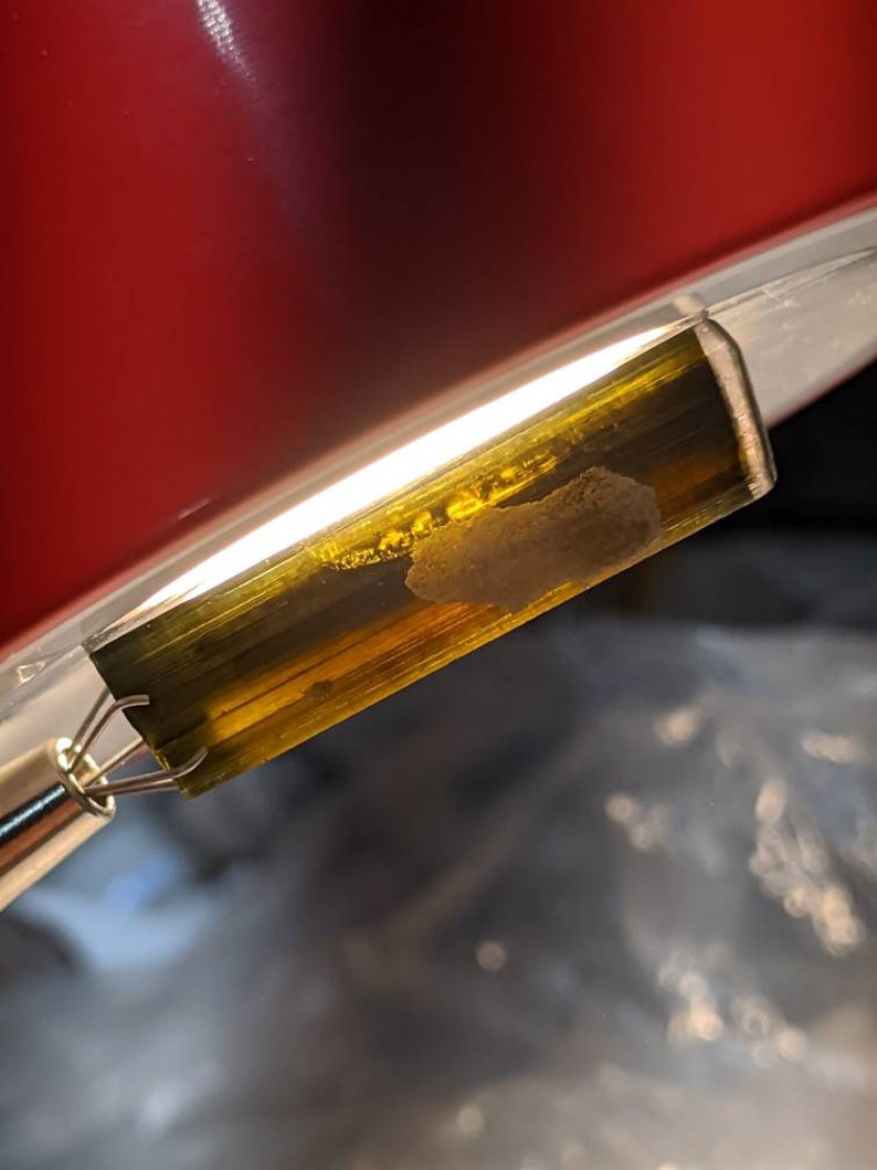 May include: A close-up of a green and gold gemstone with a white inclusion. The gemstone is held in a metal clamp.