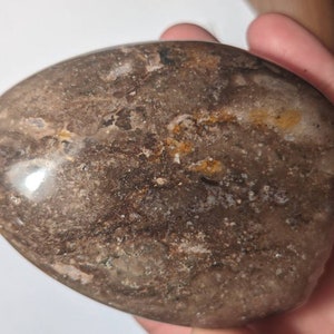 May include: A smooth, polished, brown and white agate stone with a heart shape. The stone has a natural, organic pattern with swirls and streaks of color.