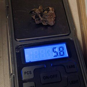 May include: A small, brown and black crystal cluster is weighed on a digital scale. The scale reads 5.8 grams.