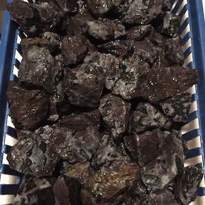 Könnte beinhalten: Ein blauer Plastikkorb gefüllt mit zahlreichen dunklen, rau strukturierten Steinen. Die Steine sind hauptsächlich dunkelbraun und schwarz, mit grünen und weißen Flecken. Die Steine scheinen nass zu sein und haben eine glänzende Oberfläche. Die Steine sind ein Naturprodukt.