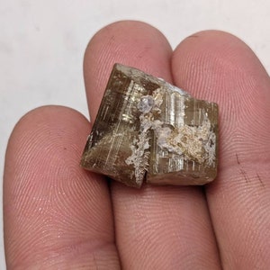 Może przedstawiać: Brązowy i biały kryształ o powierzchni prążkowanej. Kryształ ma około 5 cm długości i 2,5 cm szerokości.