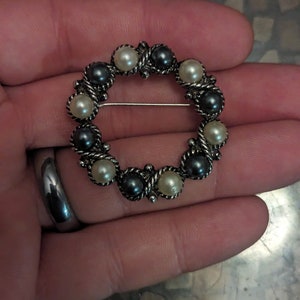 May include: A silver circular brooch with alternating black and white pearls. The brooch is made of metal and has a pin on the back.