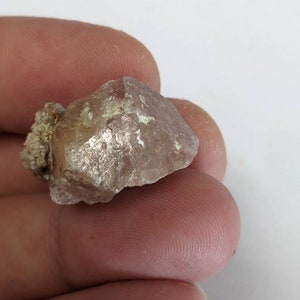 Pode incluir: Um cristal rosa e branco com uma textura áspera e natural. O cristal tem uma aparência ligeiramente translúcida e uma forma irregular.
