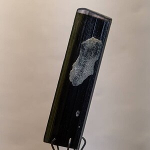 May include: A dark green and black gemstone with a rough, rectangular shape. The gemstone is held in a metal clamp.