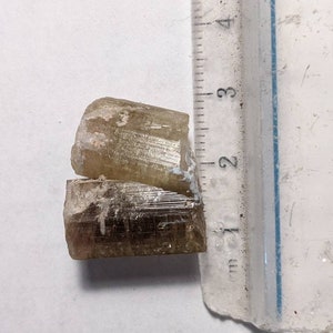 Może przedstawiać: Dwa ułożone na sobie, jasnobrązowe, przezroczyste, wydłużone kryształy z prążkami. Kryształy mają około 2 cm długości.