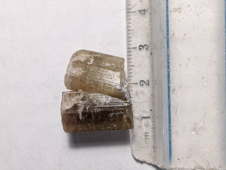 Może przedstawiać: Dwa ułożone na sobie, jasnobrązowe, przezroczyste, wydłużone kryształy z prążkami. Kryształy mają około 2 cm długości.