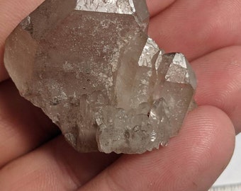 May include: A cluster of clear quartz crystals with a smoky grey hue. The crystals are clustered together in a natural formation, with some points facing upwards and others facing downwards. The crystals are translucent and have a slight sparkle.